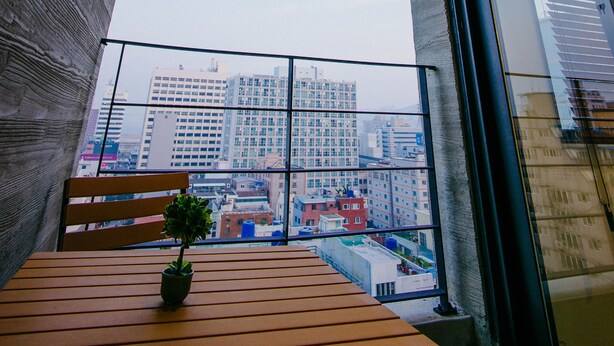 A balcony with a wooden table, a potted plant, and a city view. - Morning Dew Guesthouse (Busan)