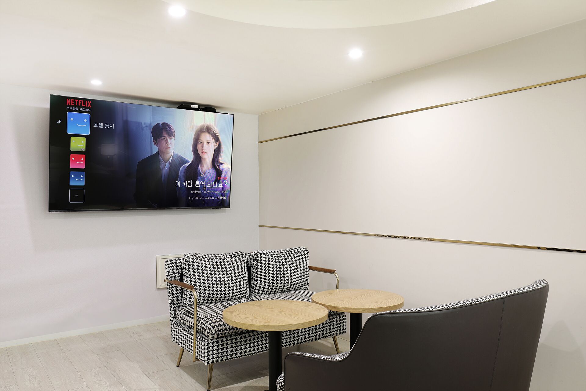 Quarto moderno com televisão de tela plana na parede exibindo um filme. Há uma poltrona com padrão xadrez de dentes de cão e uma mesa redonda pequena.
