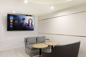 Una habitación moderna con un televisor de pantalla plana montado en la pared reproduciendo una película. Hay una silla con estampado de pied de pavo y una pequeña mesa redonda.