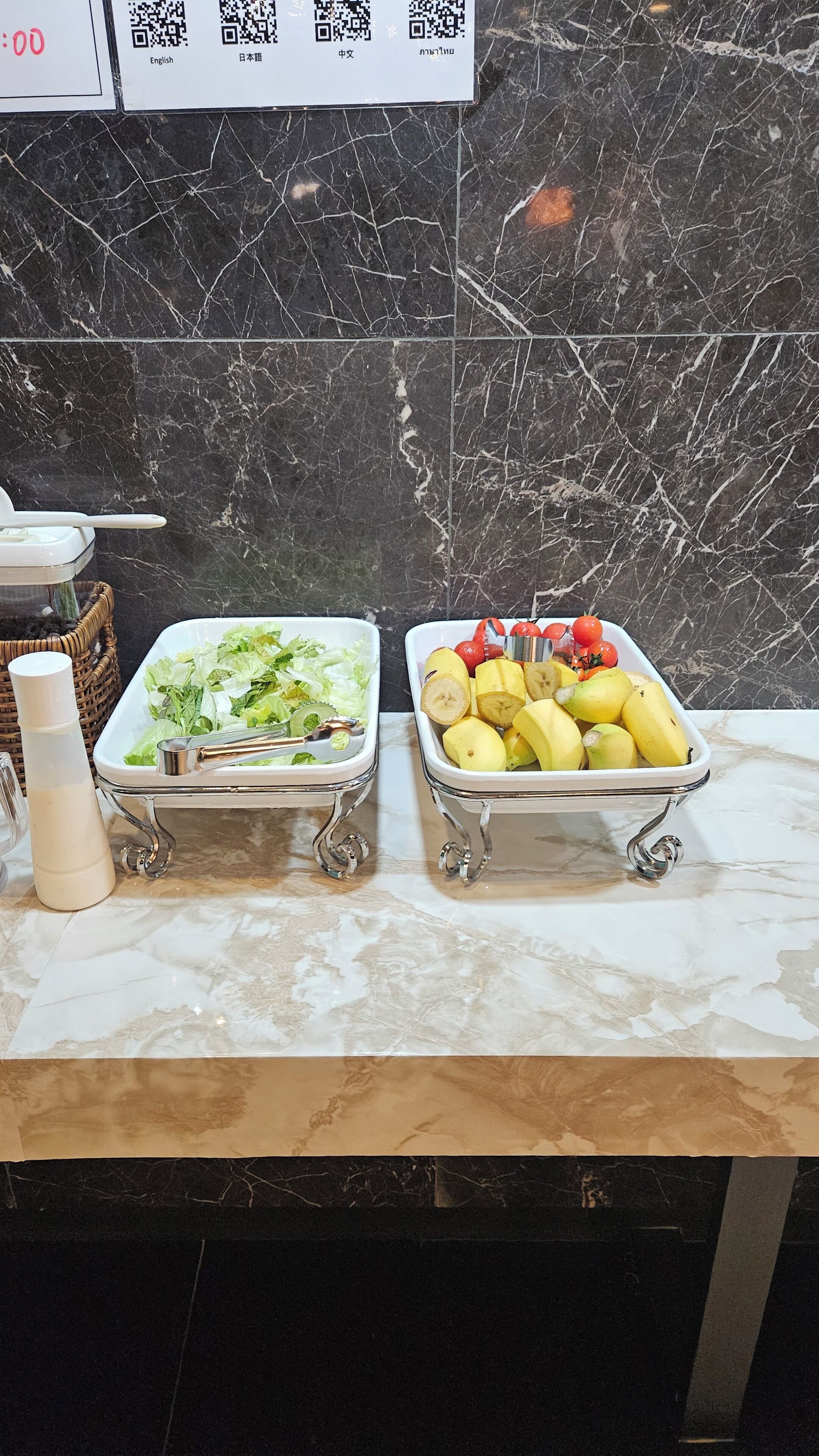 A buffet table with two trays of food items, one with sliced bananas and the other with leafy greens, on a marble countertop.