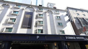 Entrada de un hotel con un llamativo letrero que dice 'Gran Inauguración', una 'P' que indica estacionamiento, y el nombre del hotel 'TOMGI HOTEL' exhibido sobre la entrada.