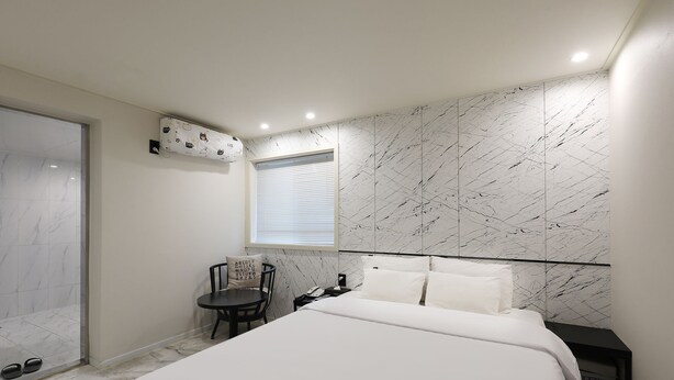 A bedroom with a bed, a chair, a small table, a nightstand, a window, and a wall with a marble-like pattern. - Hotel Tomgi Jamsil (Seoul)