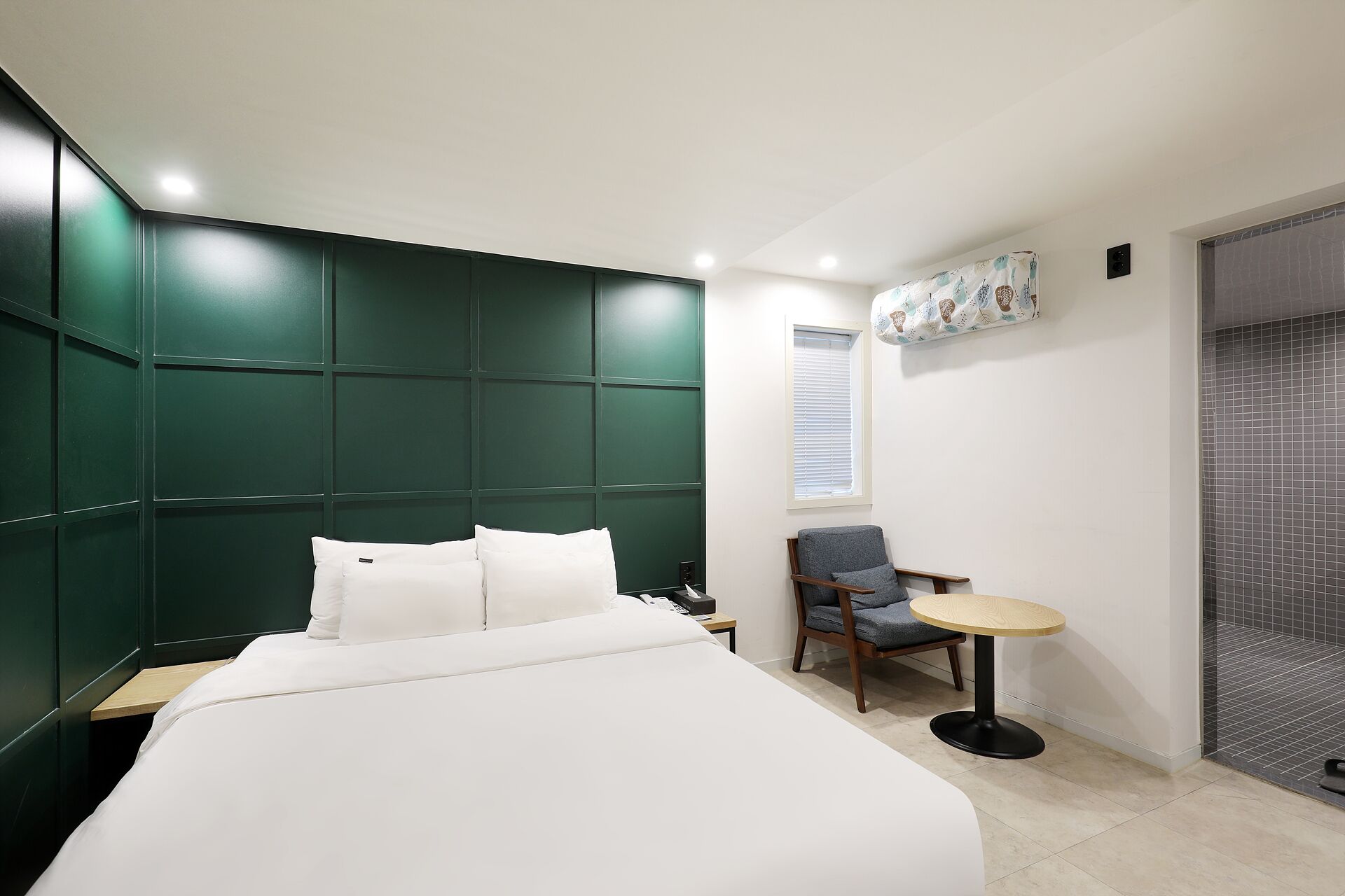 A modern hotel room with a large bed, a small table, a chair, and a window with blinds.
