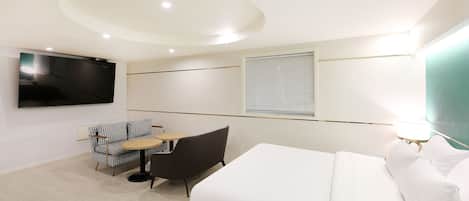 A hotel room with a bed, a TV, a sofa, a small table, and a window with blinds.
