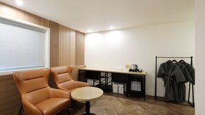 Una habitación con un sillón de cuero marrón, una mesita redonda pequeña, un escritorio de madera, un perchero y una ventana con persianas.