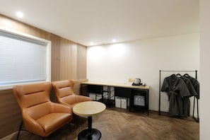 Una habitación con un sillón de cuero marrón, una mesita redonda pequeña, un escritorio de madera, un perchero y una ventana con persianas.