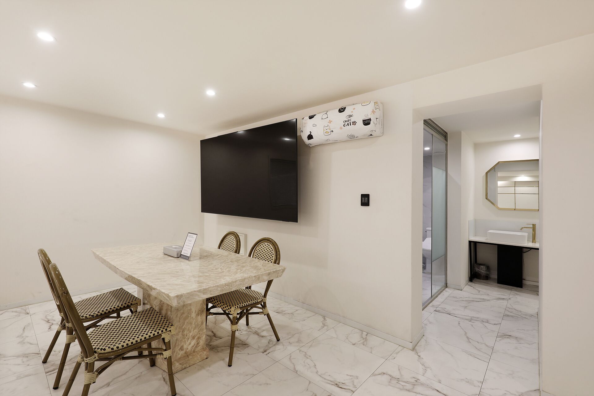 A modern dining area with a marble table, wicker chairs, a flat-screen TV, and a patterned wall hanging.