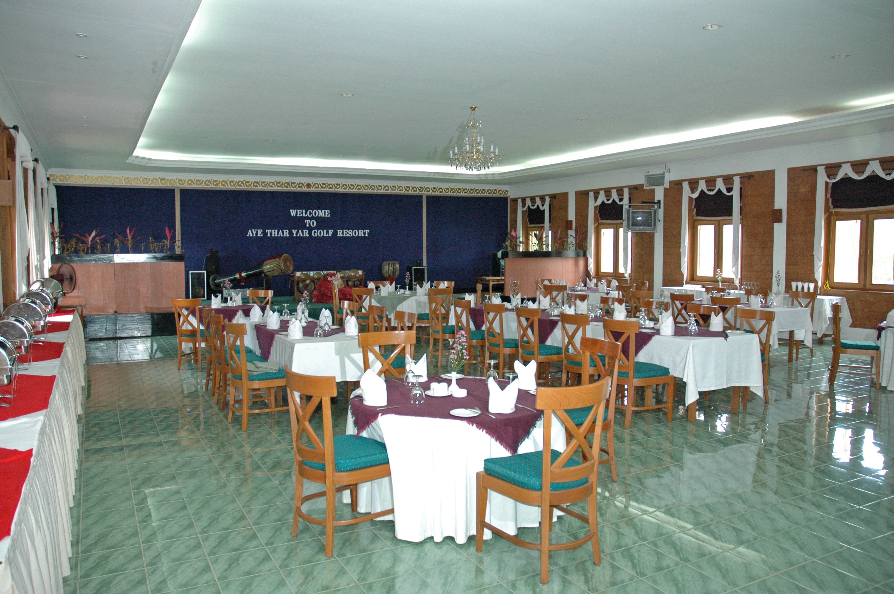 A large banquet hall with multiple tables set for dining, a stage with a backdrop, and a chandelier.
