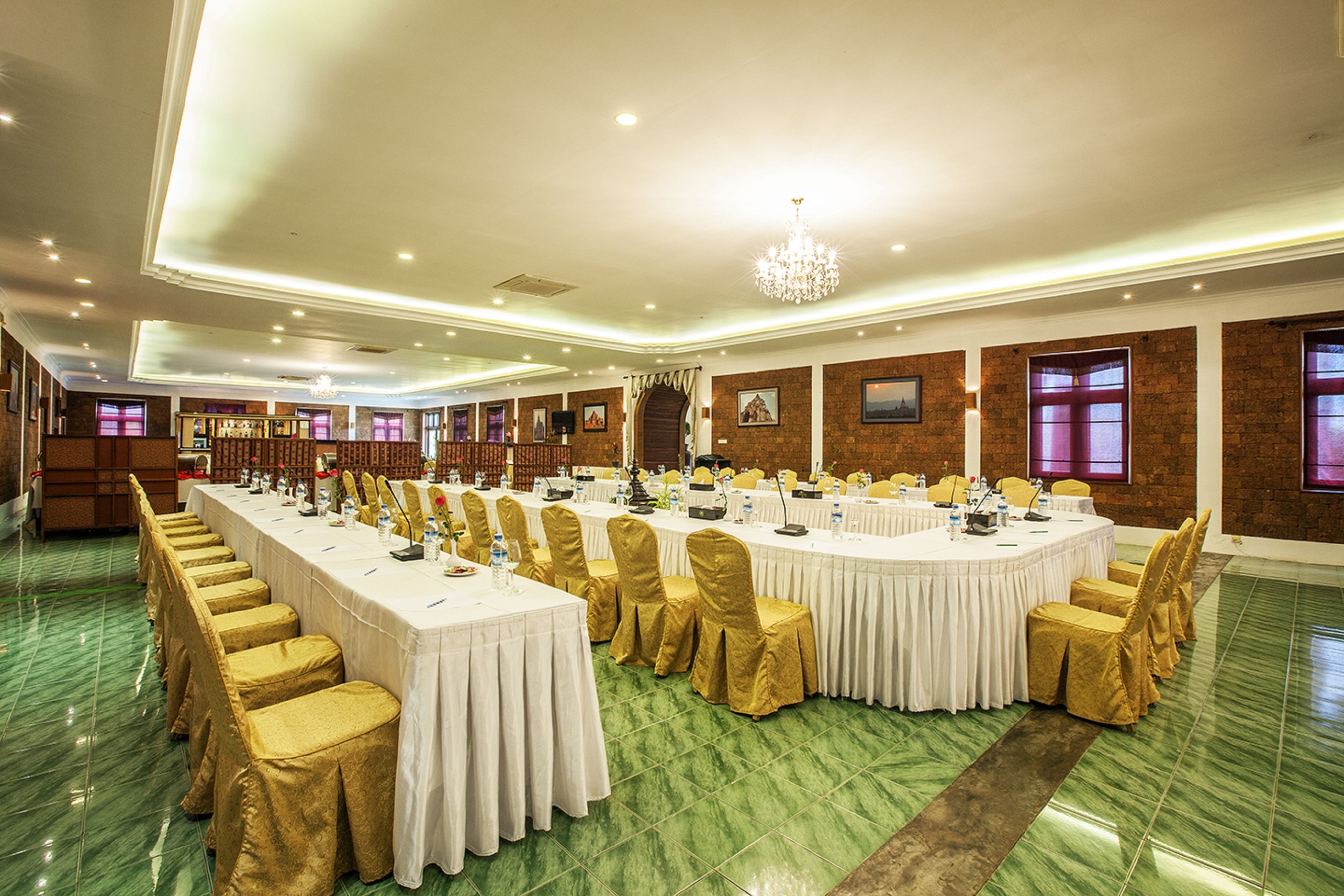 A banquet hall with multiple round tables, each surrounded by chairs with gold covers. The room features green marble flooring, brick walls, and a chandelier.