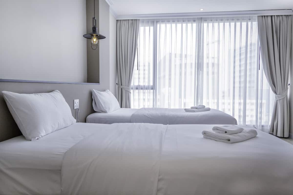 A hotel room with two beds, white linens, a bedside table, a lamp, and a window with curtains.