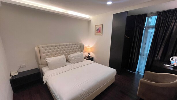 A hotel room with a large bed, a bedside table with a lamp, a chair, and a window with curtains. - Capital O 91074 Grand World Hotel (Masai)