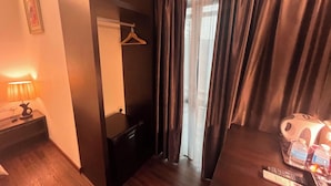 A hotel room with a wooden floor, a bedside table with a lamp, a hanging coat rack, a mirror, and a window with curtains. - Capital O 91074 Grand World Hotel (Masai)