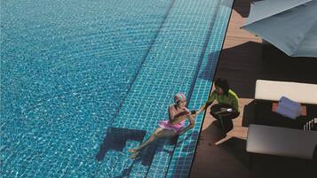 Une piscine à l’eau d’un bleu cristallin, une personne assise au bord, des chaises longues et des parasols sur la terrasse.