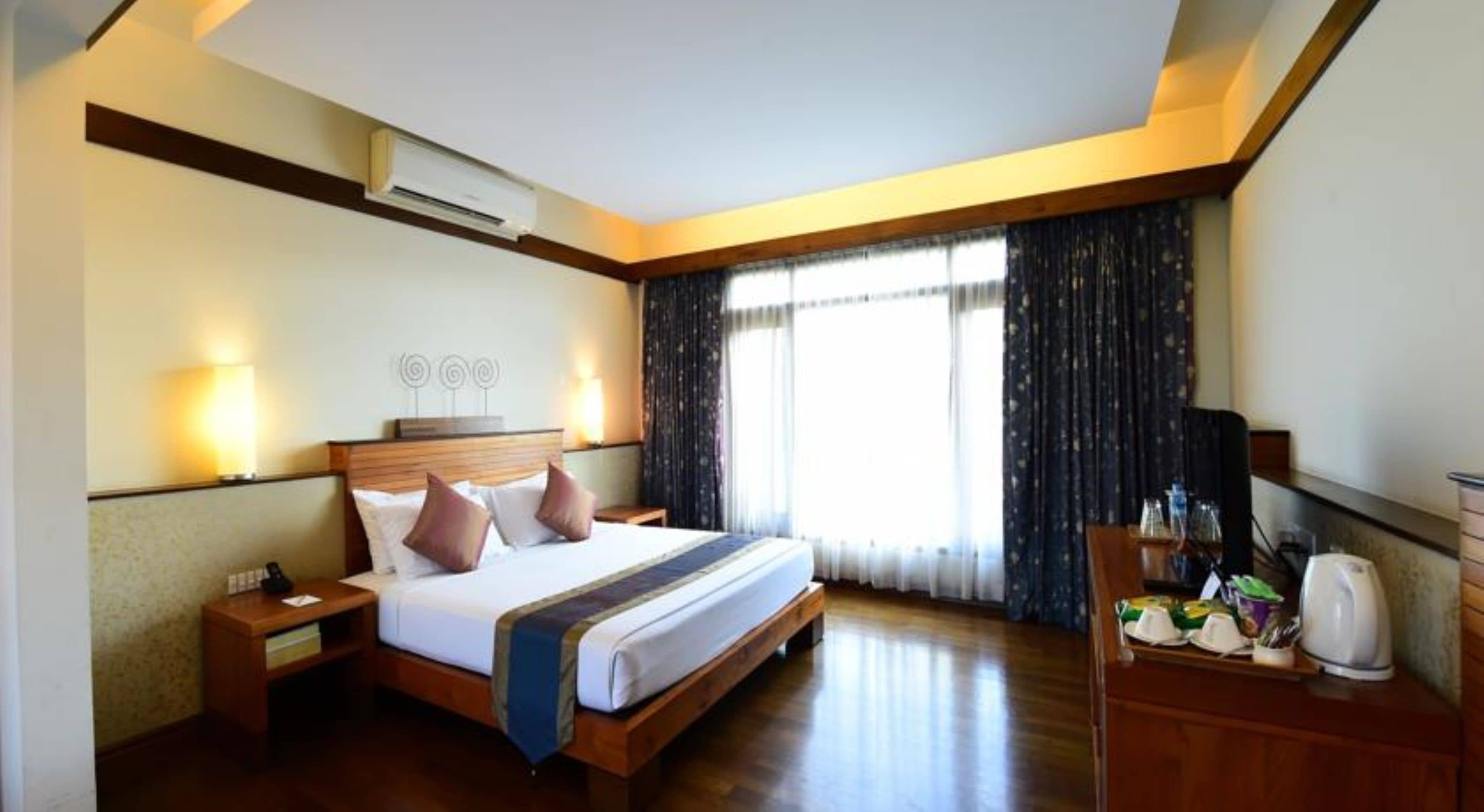 Habitación de hotel con una cama de madera, una mesita de noche, un escritorio con hervidor y un gran ventanal con cortinas.