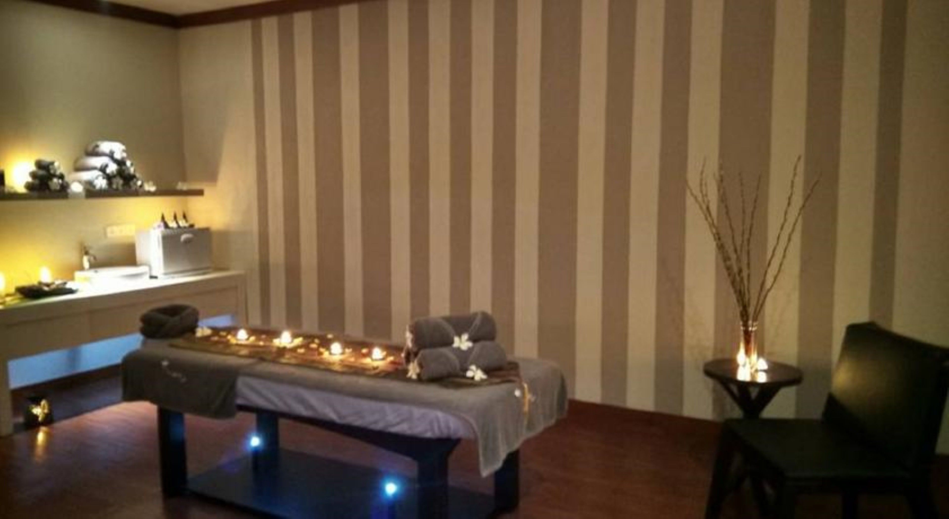 A room with a massage table, candles, a chair, and a small table with a vase.
