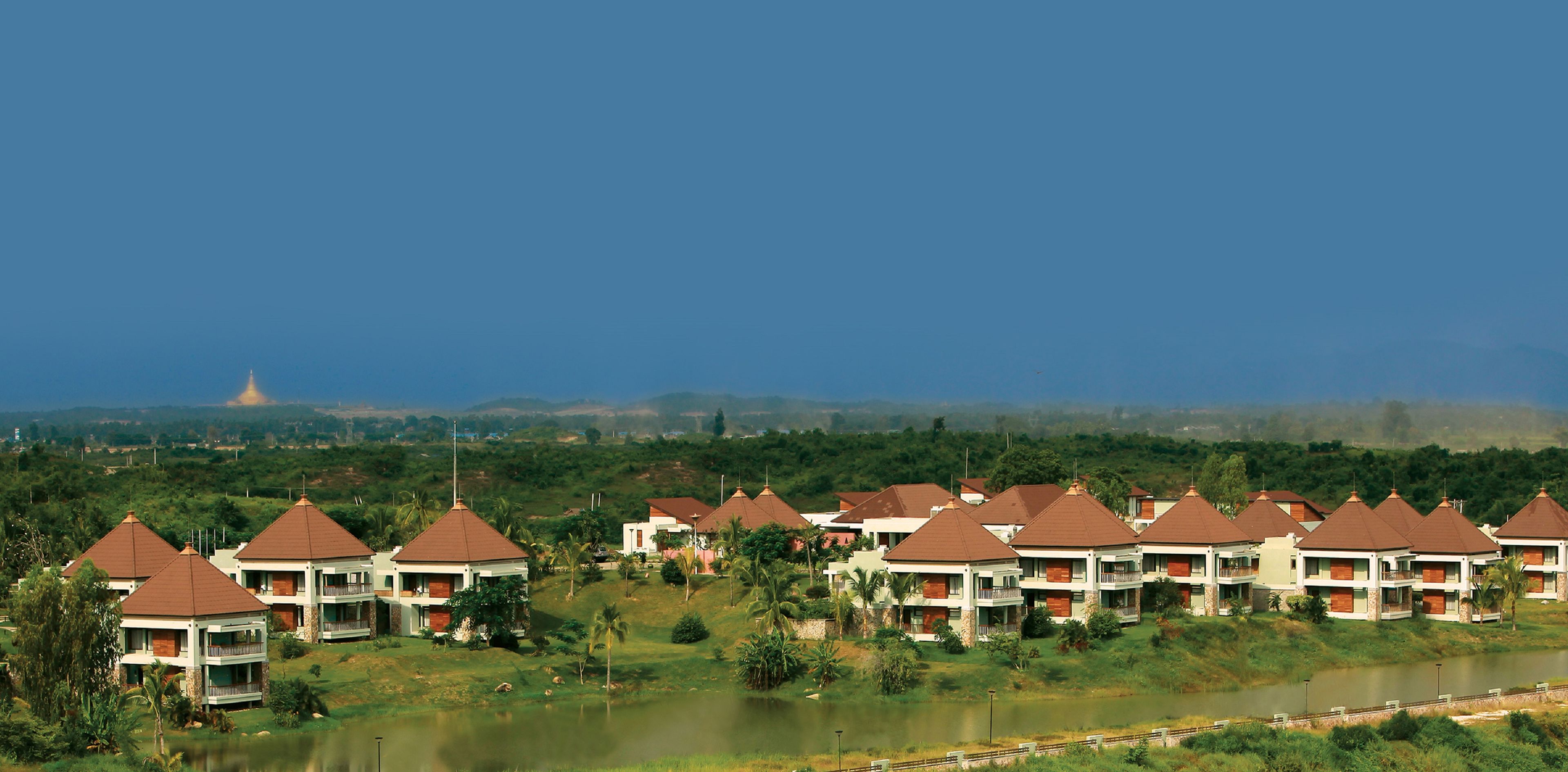 A resort with multiple bungalow-style buildings, each with a red roof and white walls, surrounded by lush greenery and a body of water.