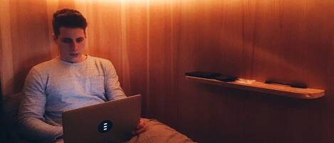 Una persona sentada en una cama con una computadora portátil, en una habitación con paredes de madera y una estantería con un control remoto.