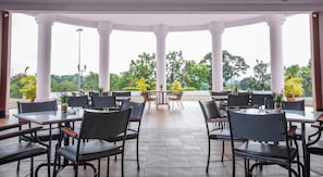 Un área de comedor al aire libre techada con varias mesas y sillas, rodeada de columnas blancas y con vistas a un jardín ajardinado.