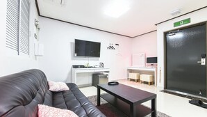 A modern hotel room with a black leather sofa, a small wooden table, a flat-screen TV, a desk with a computer, and a black door with an emergency exit sign. - Itaewon A One Hotel (Seoul)