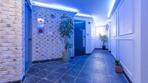 A hallway with a brick wall, a potted plant, a door with a red emergency button, and a blue-lit ceiling. - Itaewon A One Hotel (Seoul)
