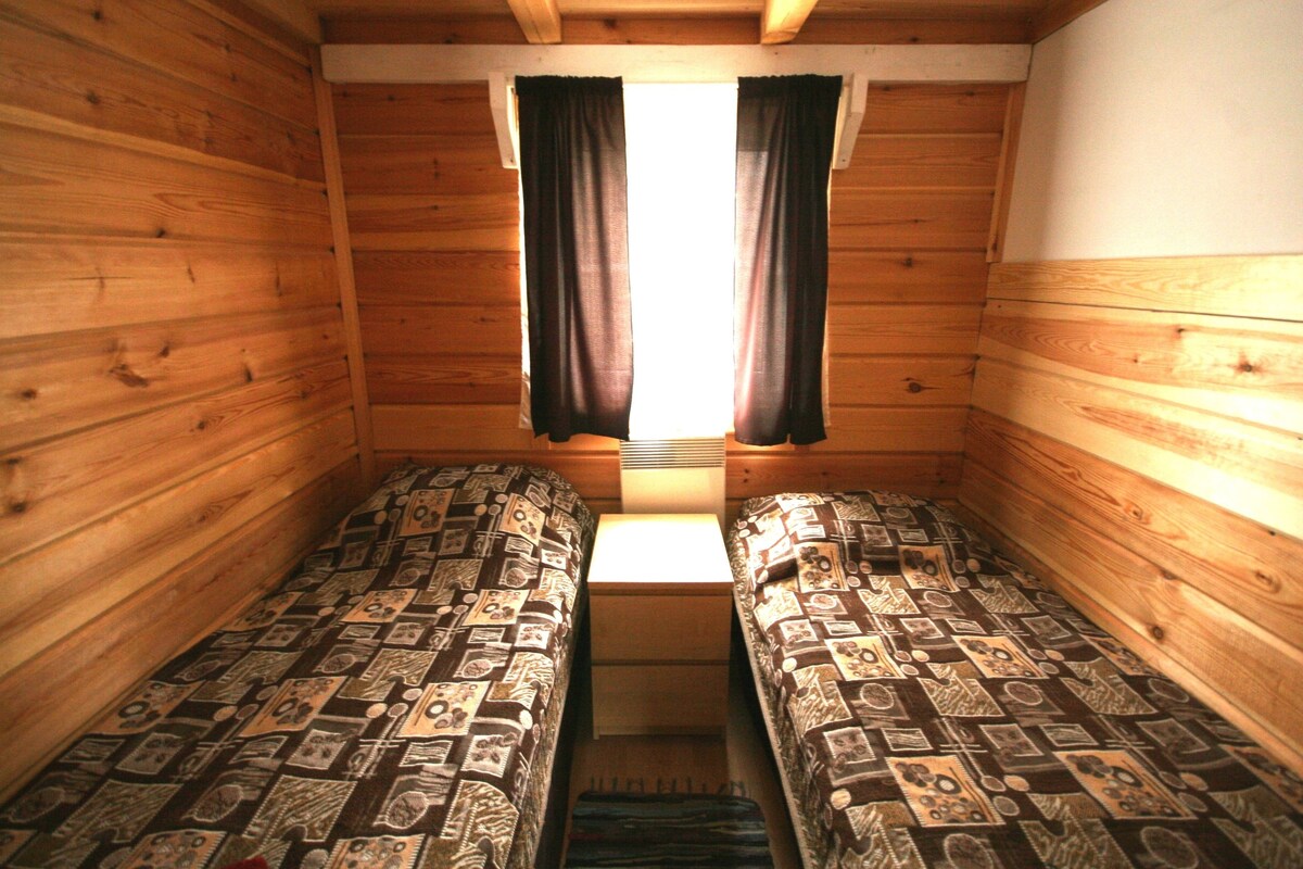 Una cabaña con dos camas de madera, una ventana con cortinas y una pequeña mesa en el centro.