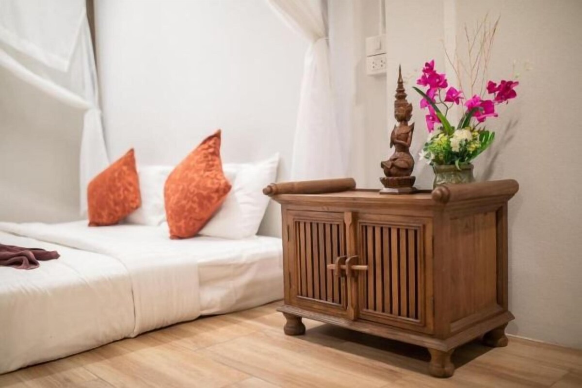 A bedroom with a white bed, orange pillows, a wooden nightstand, and a vase with flowers.