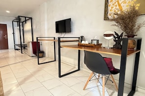 Une chambre moderne avec un bureau en bois, une chaise noire, un téléviseur fixé au mur et une plante décorative.