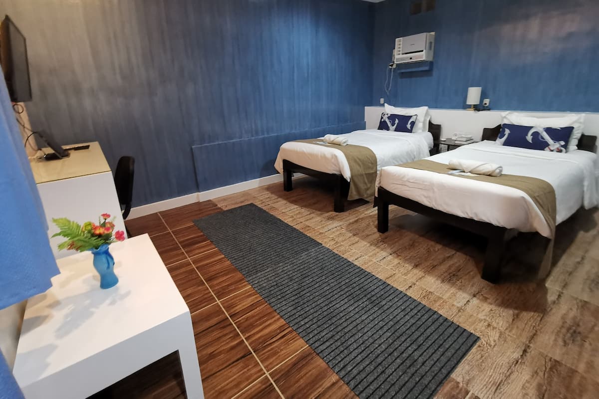 A hotel room with two beds, a desk, a chair, a small table with a vase, and a wall-mounted air conditioner.
