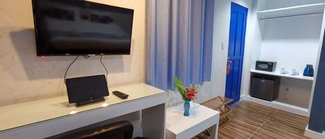 A hotel room with a flat-screen TV mounted on the wall, a small table with a vase of flowers, a radiator, and a waste bin.