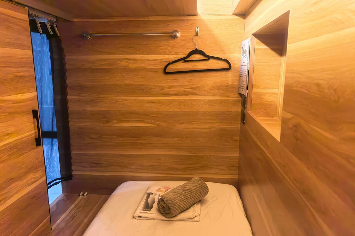 A compact room with wooden walls, a bed with a rolled towel and a magazine, a hanging black hanger, and a small window with curtains.