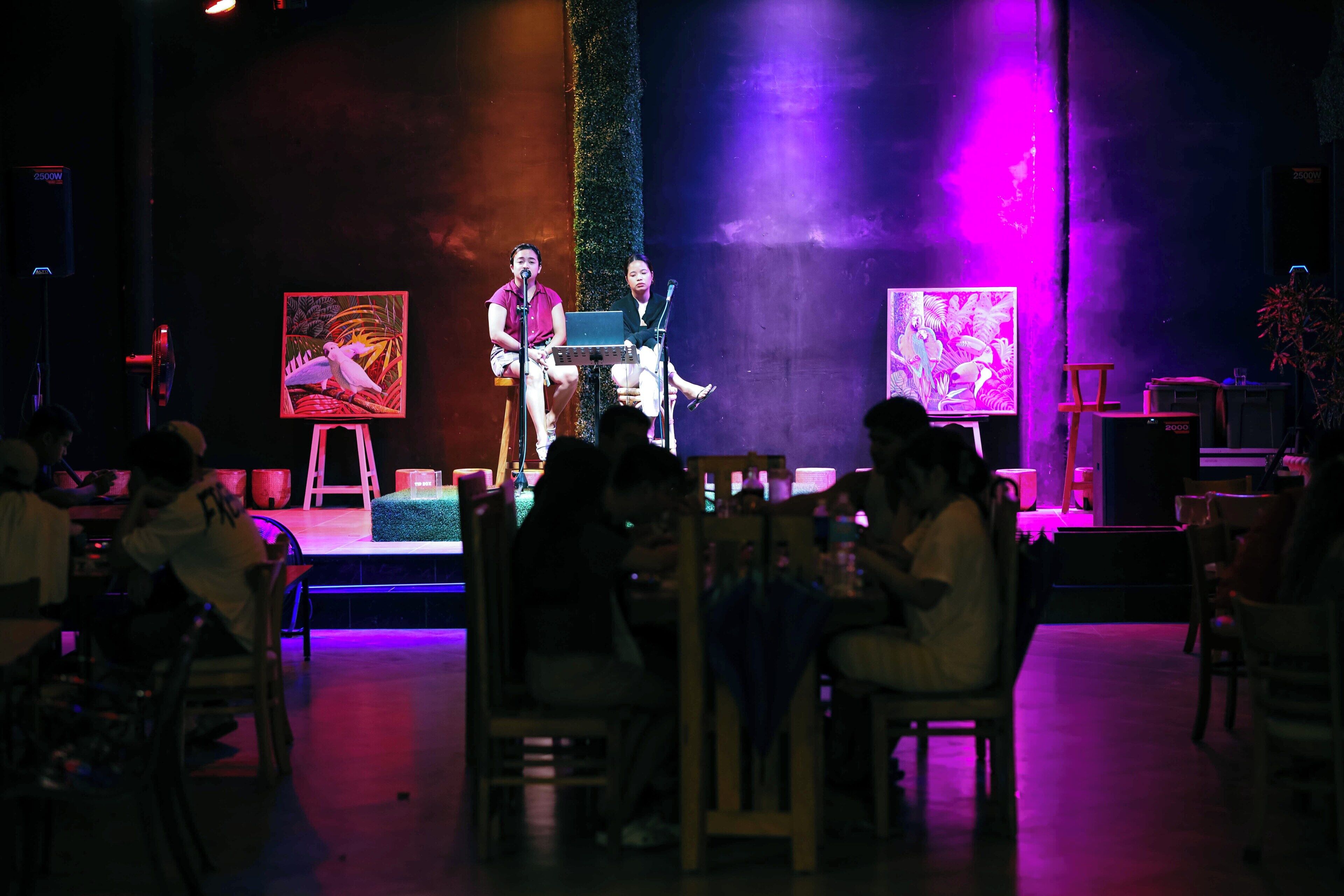 Un escenari amb dos artistes, un públic assegut i obres d'art a les parets.