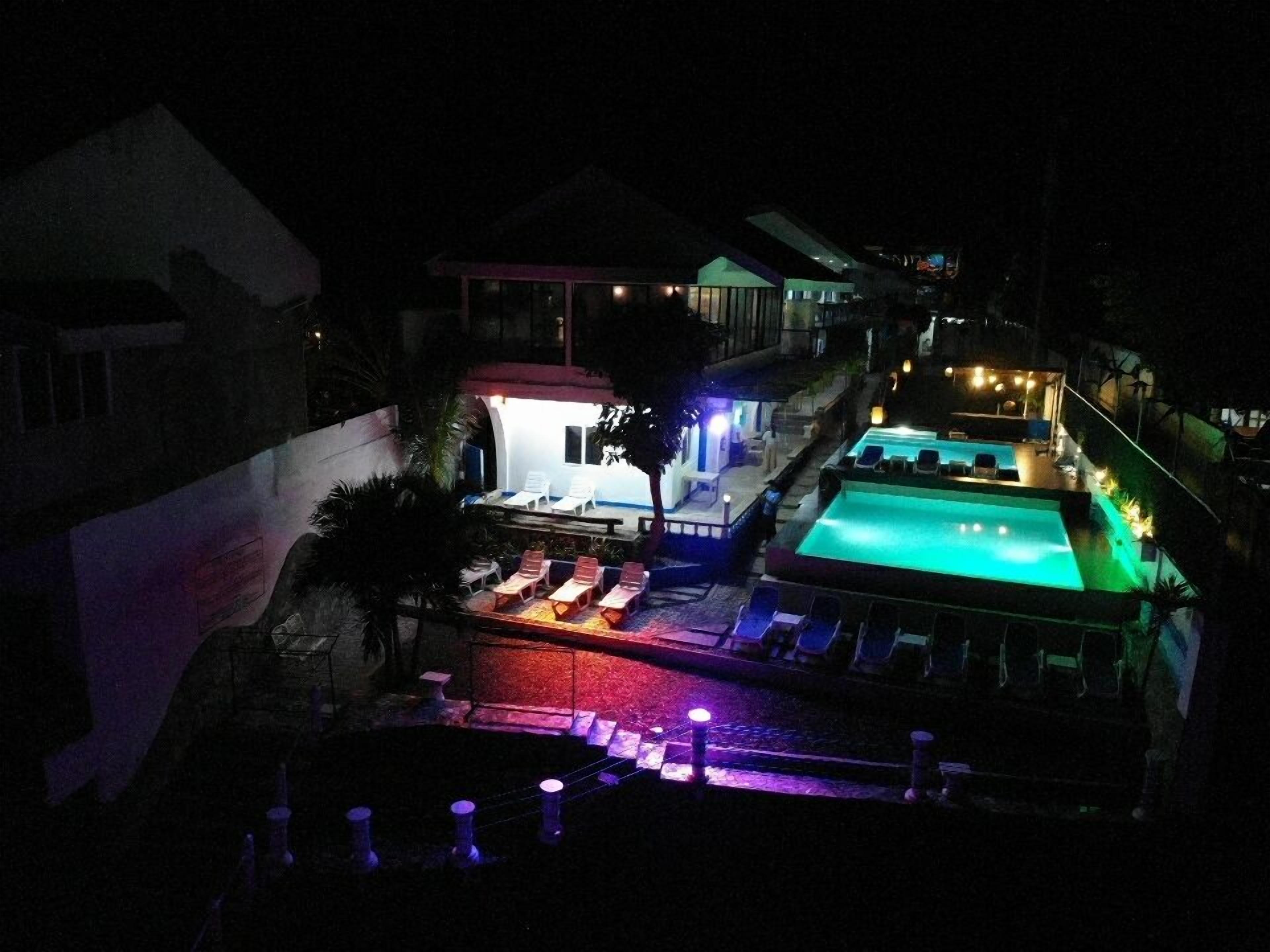 A nighttime view of a hotel with a swimming pool, lounge chairs, and a small bar area.