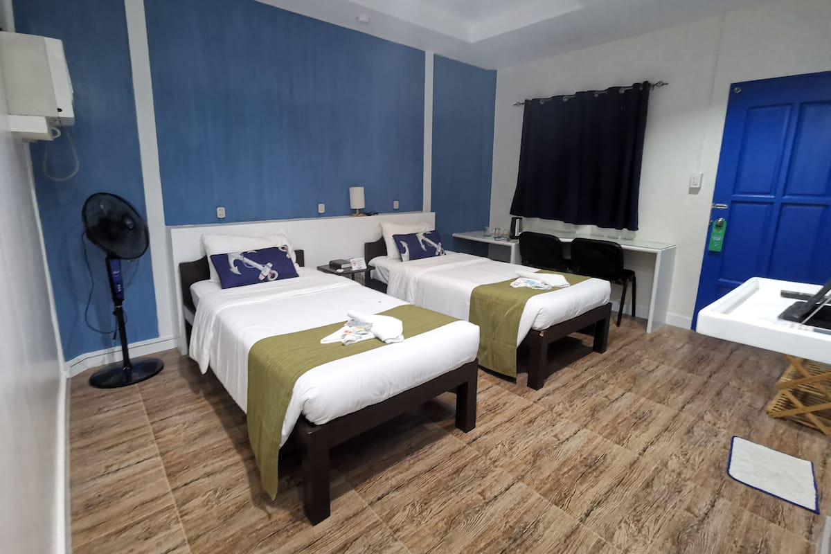A hotel room with two beds, a desk, a chair, a fan, and a blue door.