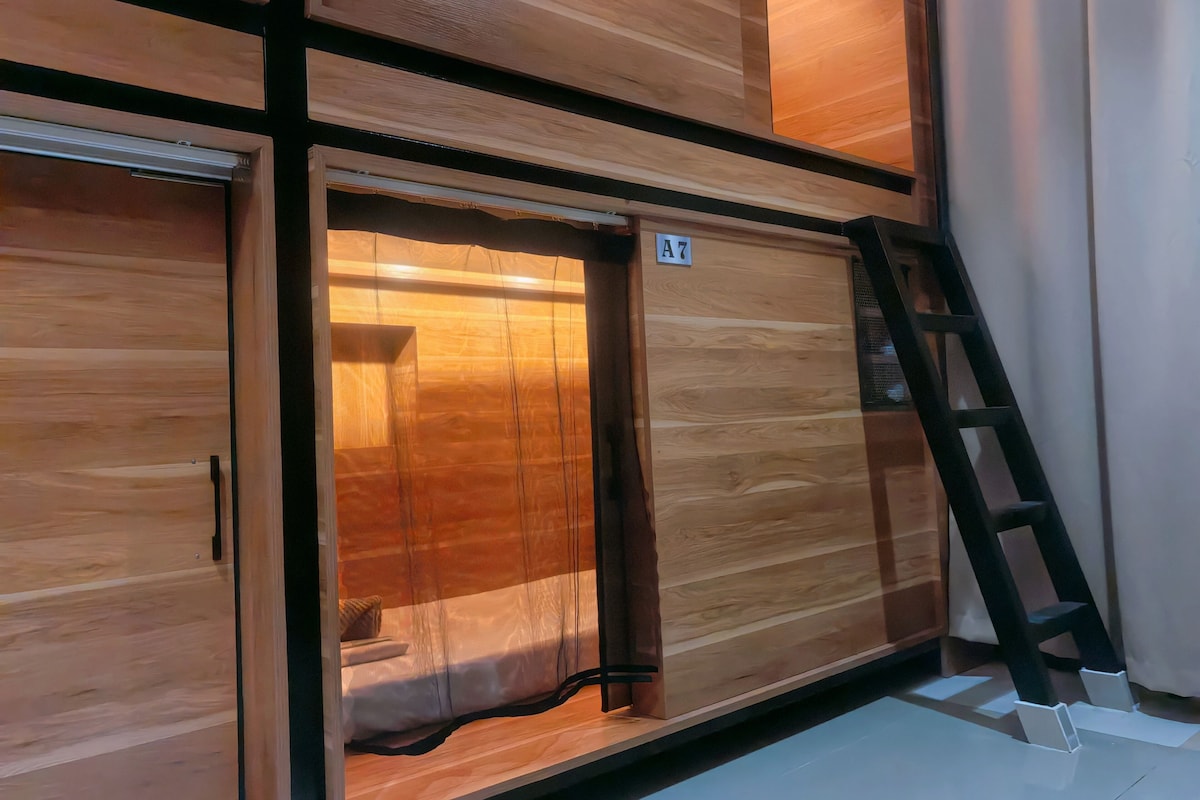 A room with a wooden floor, a bed with white bedding, a ladder, and a door marked with the number A7.