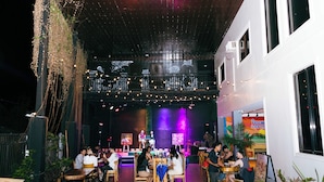 Un comedor interior con mesas y sillas, un escenario con artistas, y un balcón con luces de cuerda.