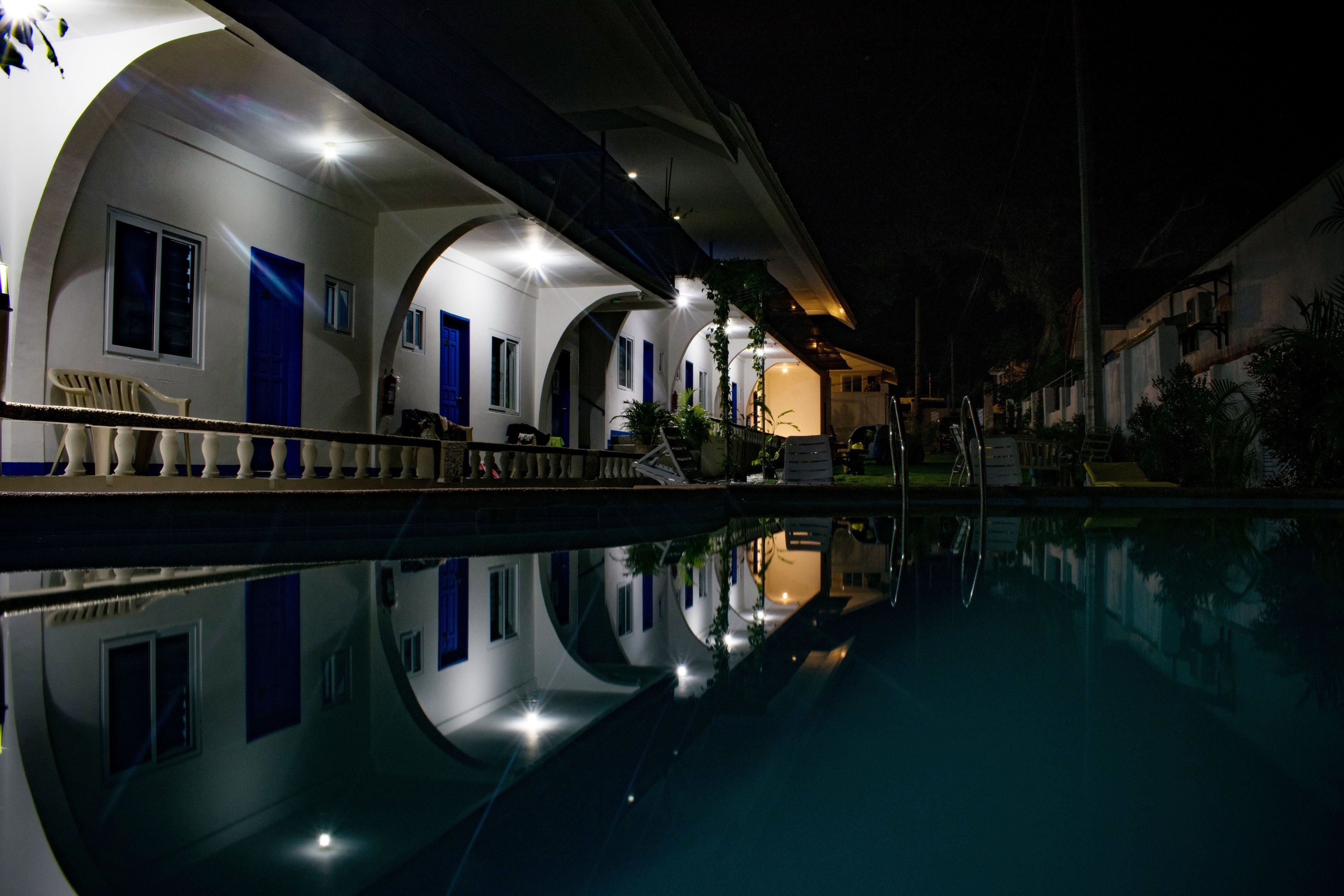 A nighttime view of a hotel with a swimming pool. The hotel features white walls with blue accents, arched windows, and a balcony overlooking the pool.