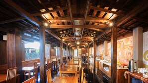 Un restaurante largo y estrecho, con vigas de madera y un techo alto. Mesas y sillas están dispuestas a lo largo de las paredes, y hay un mostrador con una cafetera y tazas.