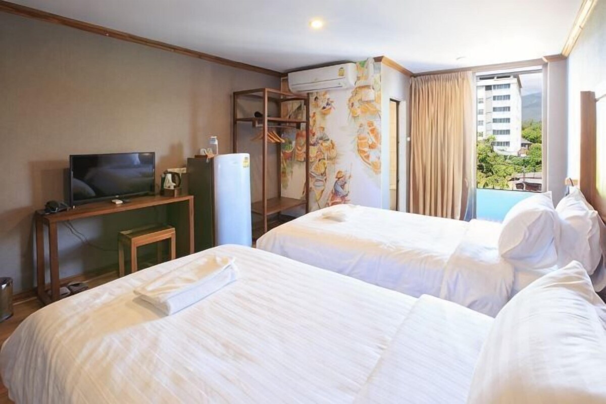 A hotel room with two beds, a television, a desk, and a window with a view of a building and greenery.