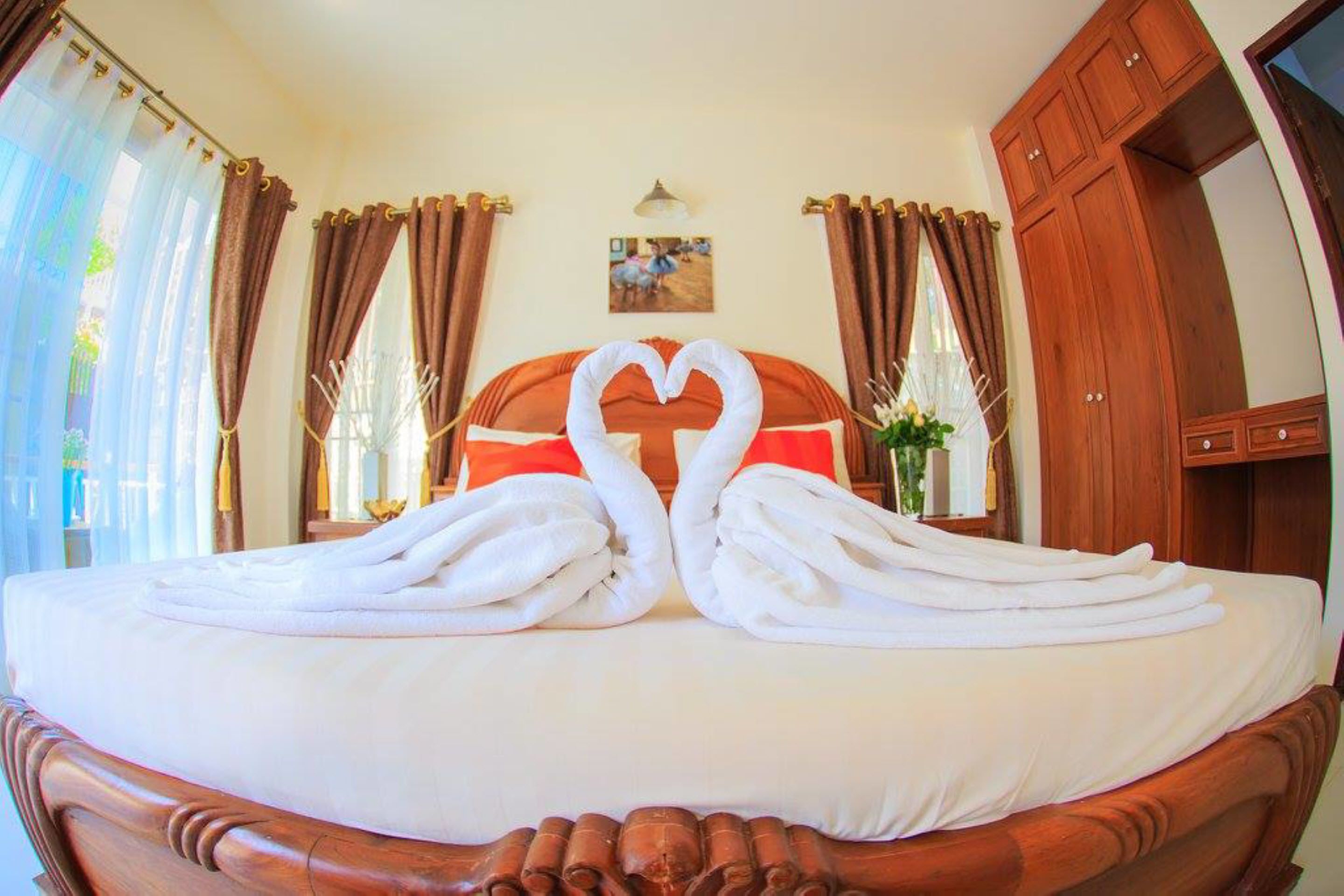 A hotel room with a bed featuring a heart-shaped towel arrangement, curtains, a painting on the wall, and wooden furniture.
