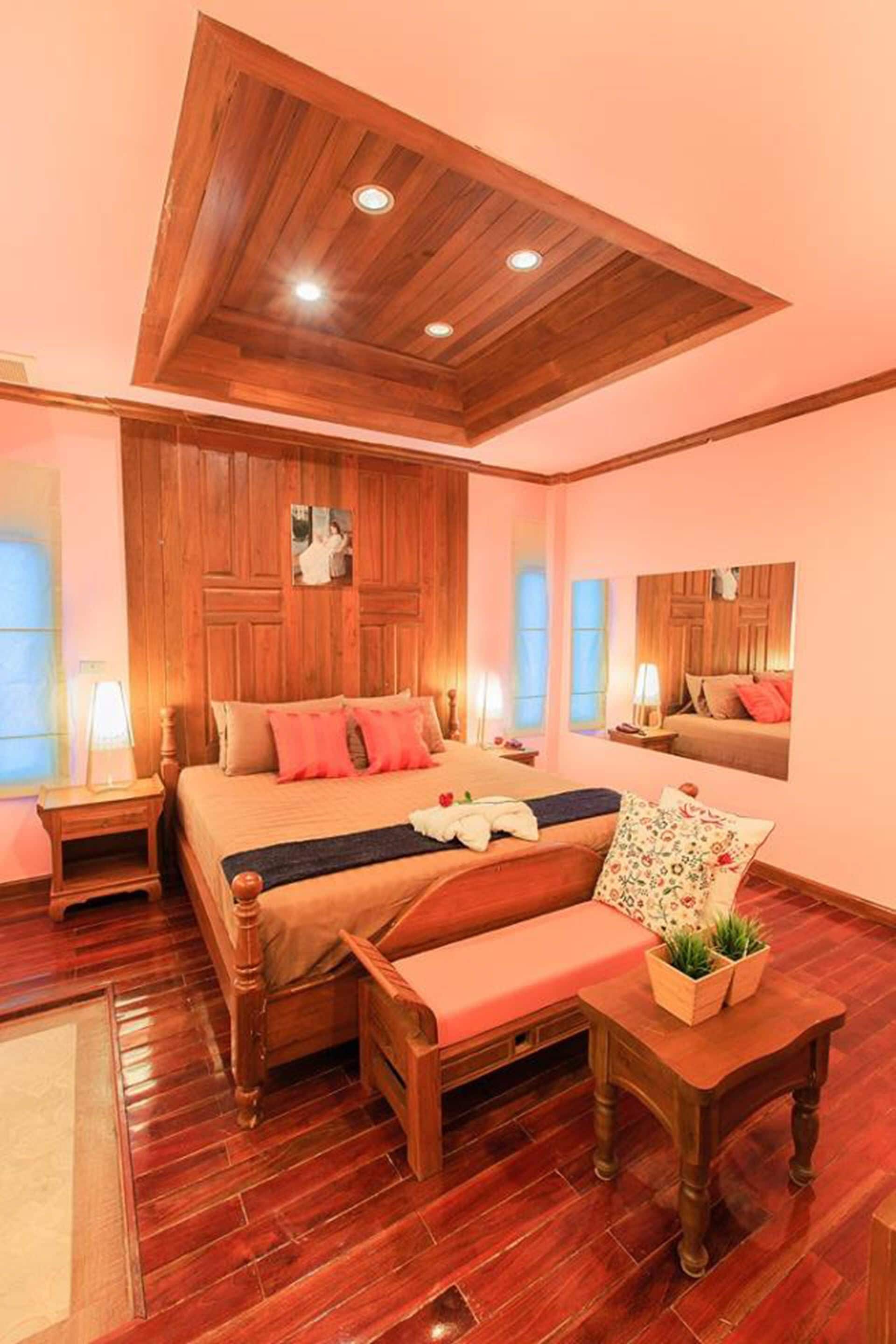 Een slaapkamer met een houten plafond, een groot bed met veel kussens, een bankje, een kleine tafel met planten, en een spiegel aan de muur.