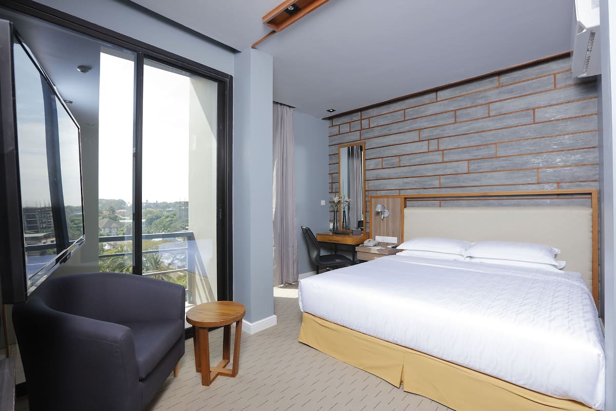 Camera d'albergo con un letto grande, un divano, un tavolino, una scrivania con sedia, uno specchio e vista sulla città attraverso ampie finestre.