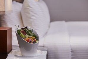 A white bowl of fruit salad on a white table next to a bed with white bedding.