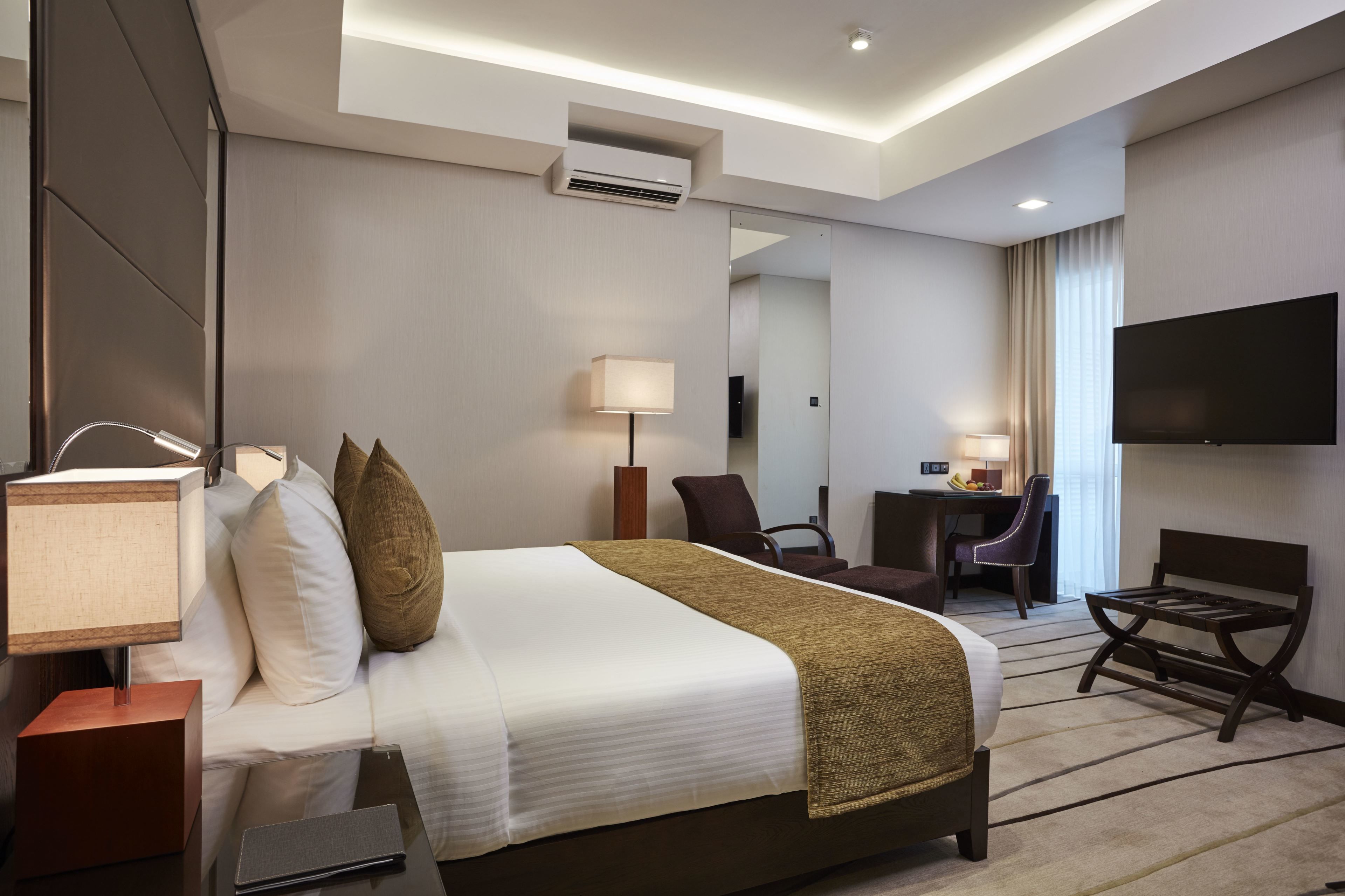 A hotel room with a large bed, bedside tables, a desk, a chair, a TV, and an air conditioning unit.