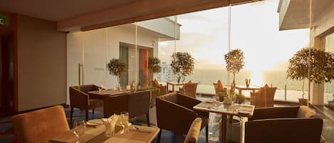 A modern restaurant with tables and chairs, large windows, and potted plants.