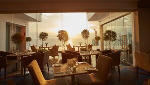 Un restaurante con mesas y sillas, ventanas amplias y plantas en macetas.