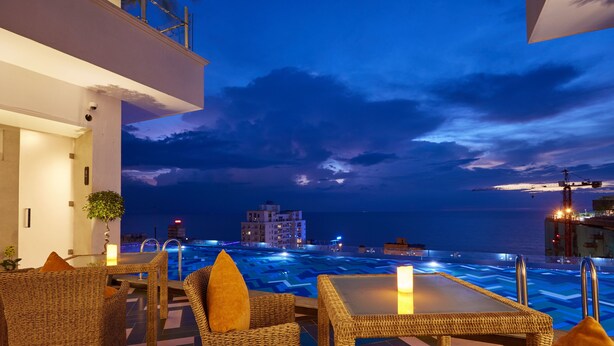 Una terraza en la azotea con mobiliario de mimbre, una piscina y vistas de la ciudad al atardecer.