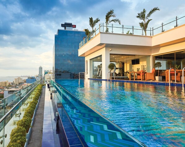 A rooftop infinity pool with a glass balustrade, a fitness area, and a seating area with outdoor furniture.