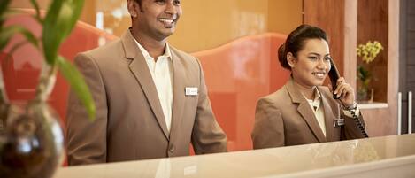 Ett receptionsområde med en receptionist bakom en glasskärm, en man som står till vänster och en kvinna som pratar i telefon till höger.