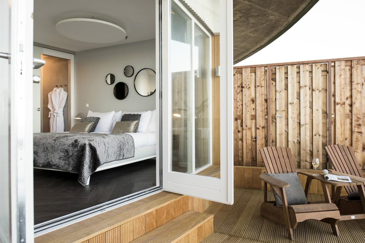 A modern bedroom with a bed, a glass door leading to a balcony, and a wooden deck with a chair and table.