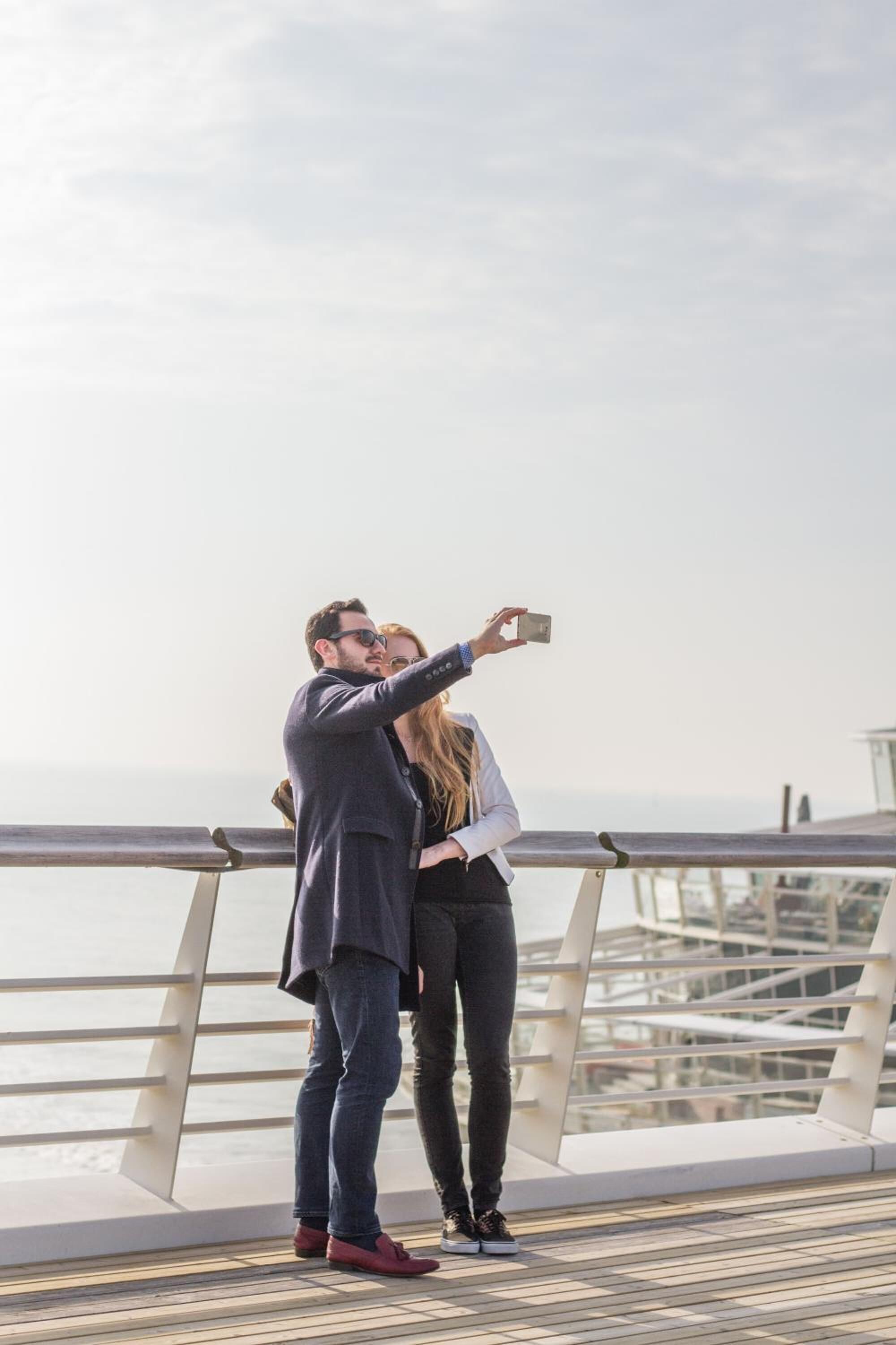 Una pareja se toma un selfie en un puente con barandilla y vista al agua.